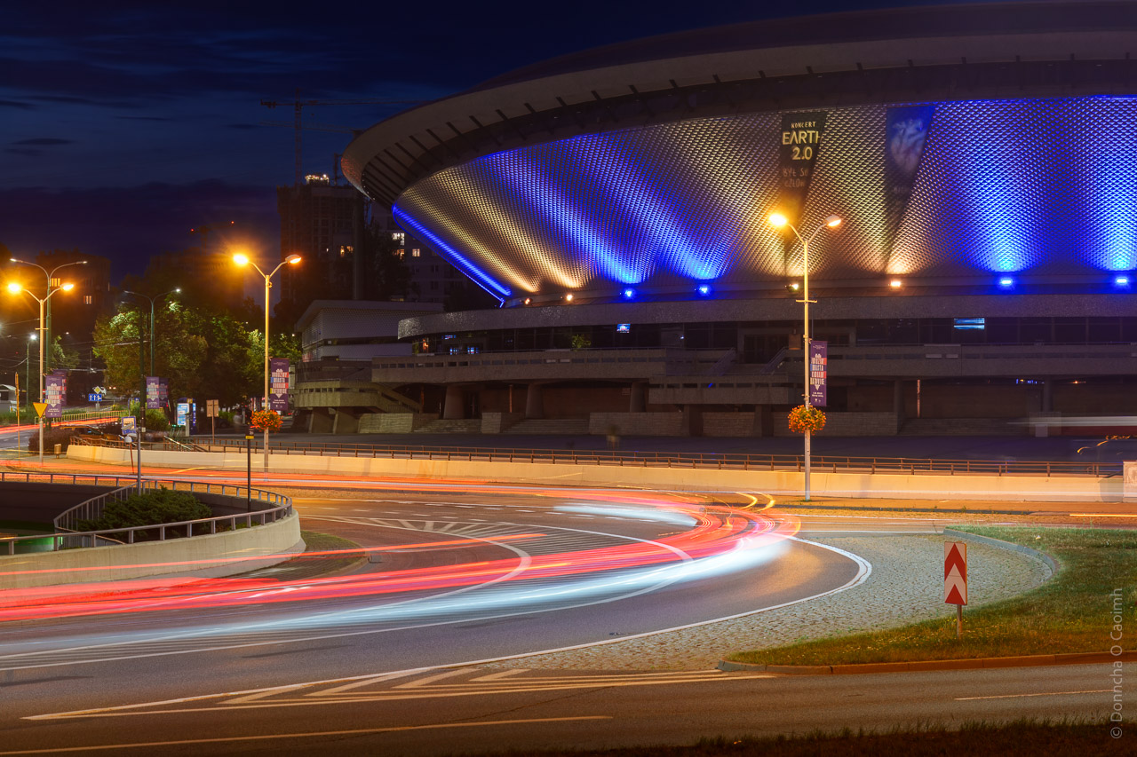 The Spodek is a large arena that is lit up at night in orange and blue lighting. It sits in front of a 3 lane roundabout and this long exposure shot captured the light trails of passing cars at night.