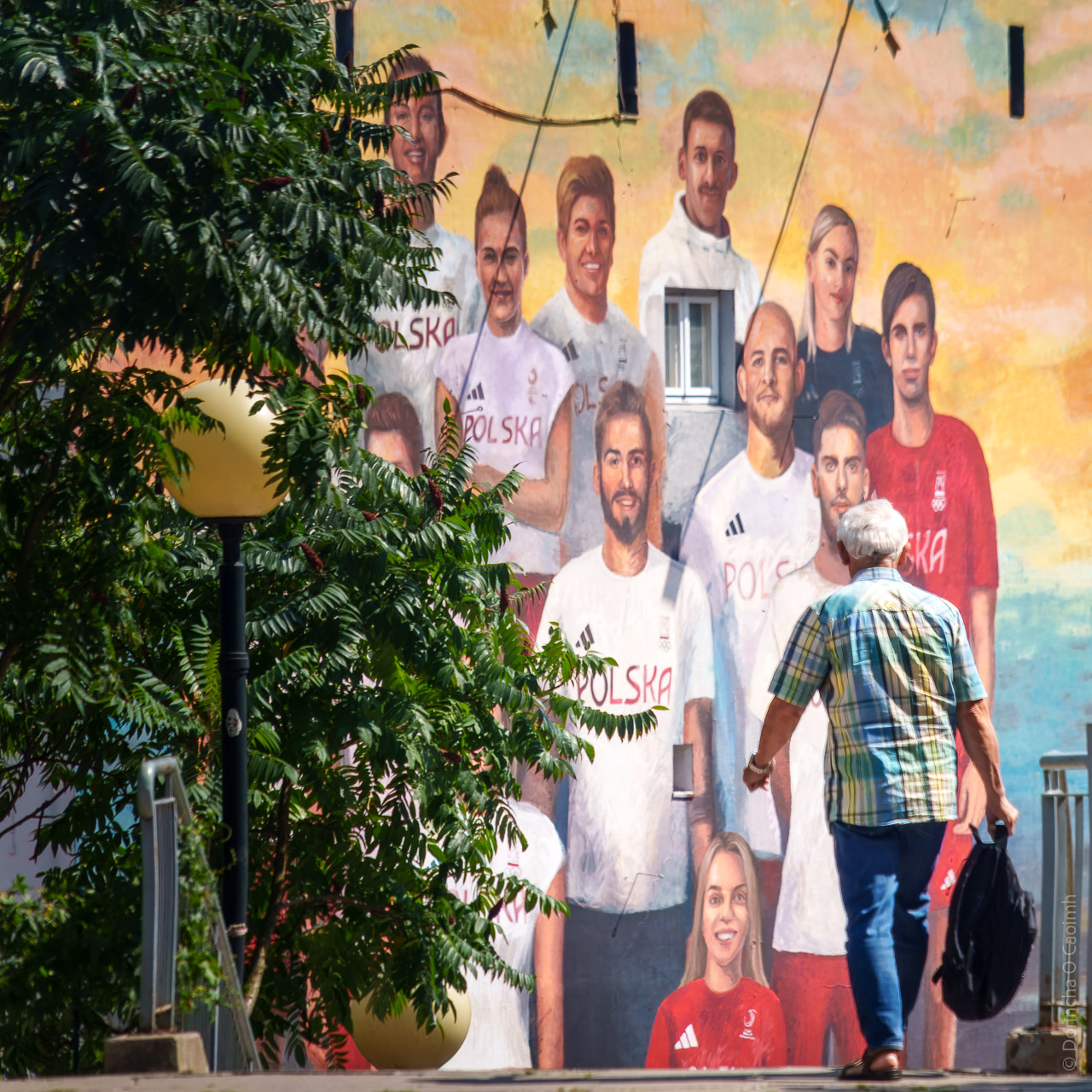 A man walks with a backpack in his hand towards stairs with a mural of Polish athletes in the background.