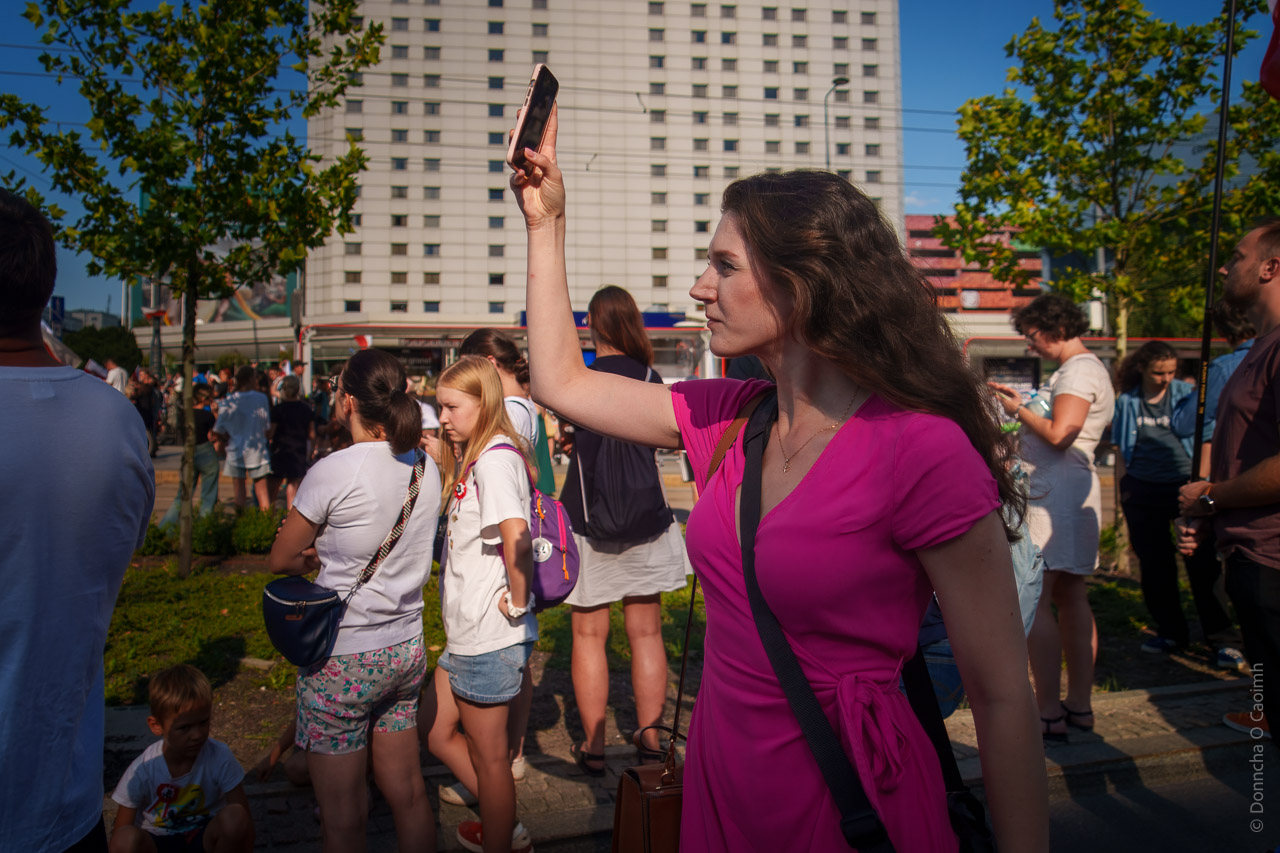 A woman in a pink dress holds up her phone on a street in Warsaw. A crowd is pictured behind her.