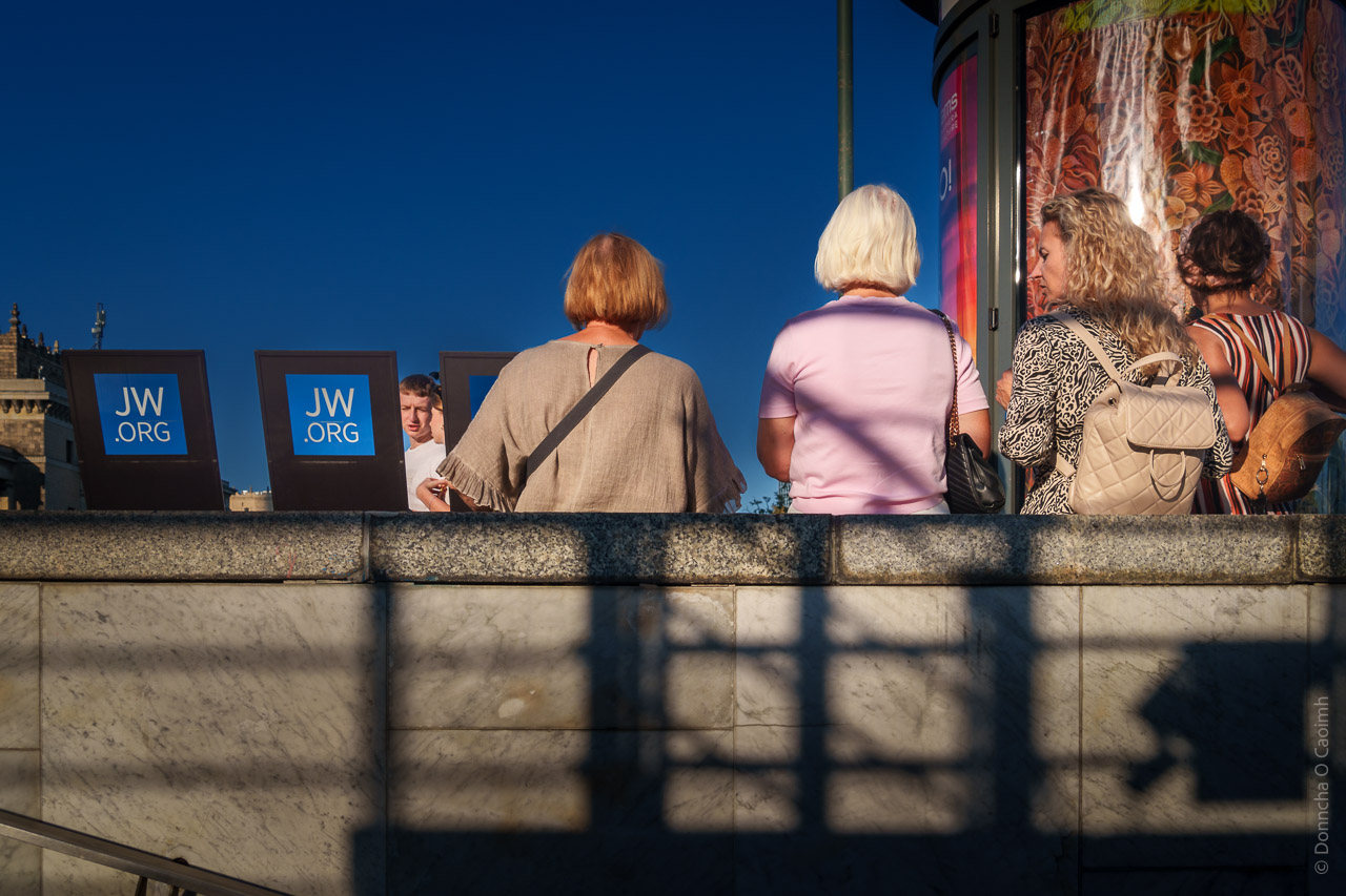 Four women lean against a wall next to their JW.ORG branded book stands that hold JW literature. A couple can be glimpsed walking by and the sky is a clear blue.