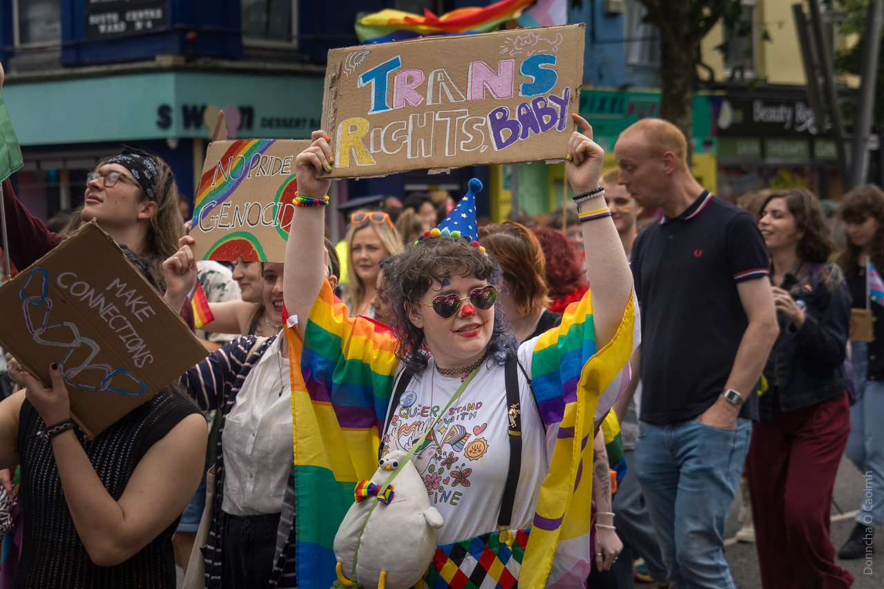 A woman holds a sign in the air saying "TRANS RIGHTS, BABY" while walking in the parade.
