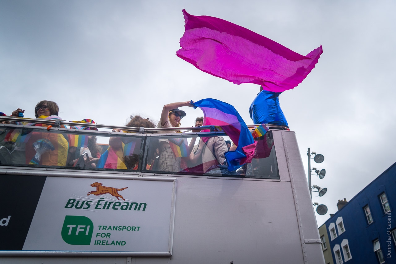 People on a bus in the parade. Someone holds the transgender flag over the side of the bus while someone else waves a bright pink flag out.