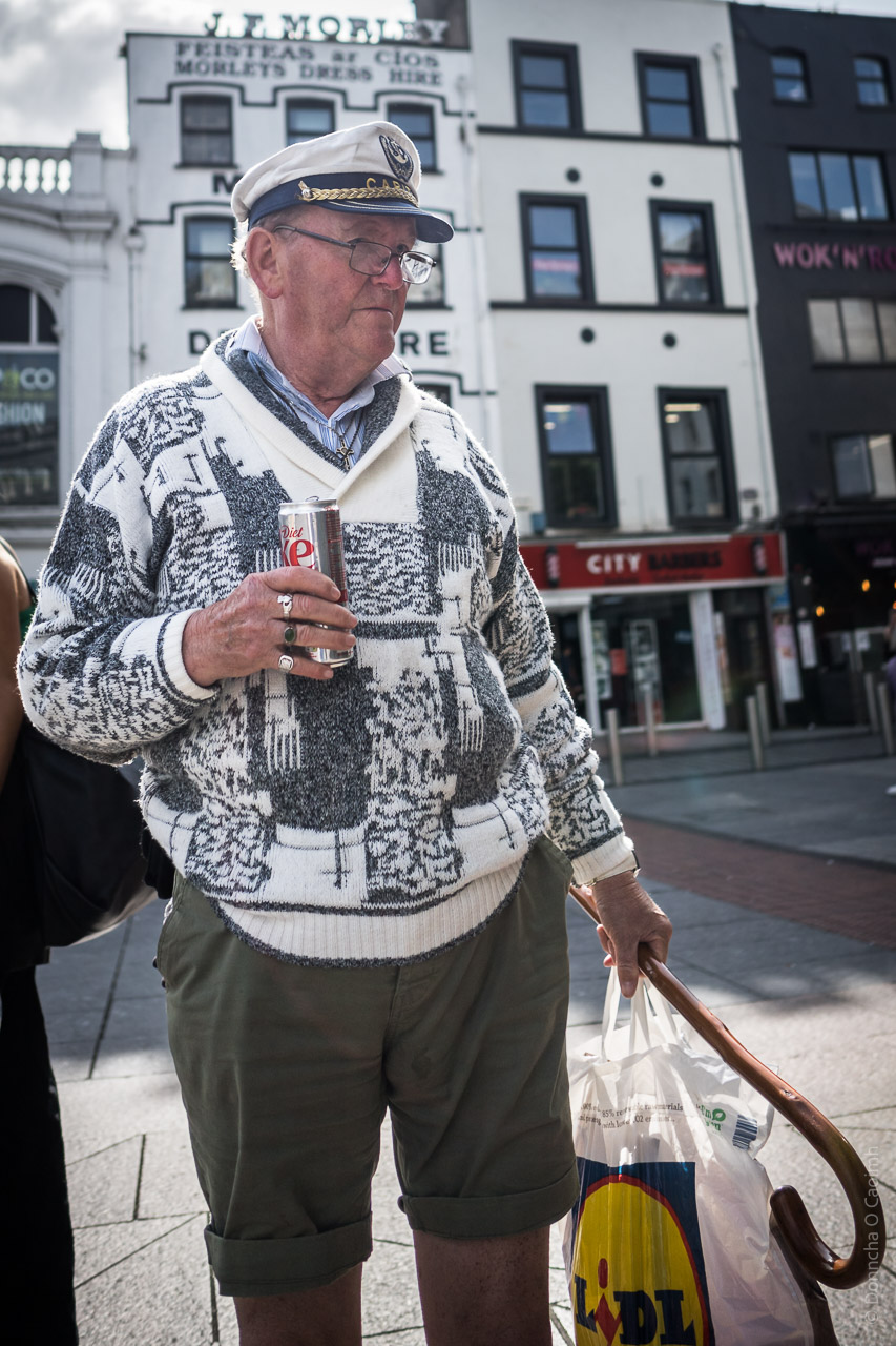 A man wearing a naval cap, with the text Capitan on it (visible from a different photo) looks to the side with a diet Coke in one hand and a shopping bag/walking stick in the other.
He's wearing shorts.