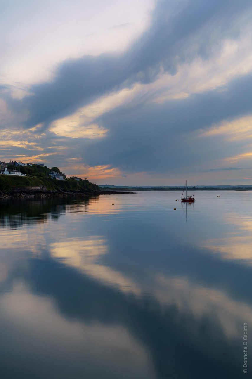 The sky is mirrored in the calm waters. The sun is setting and a boat is in the water while clouds hang in the sky.