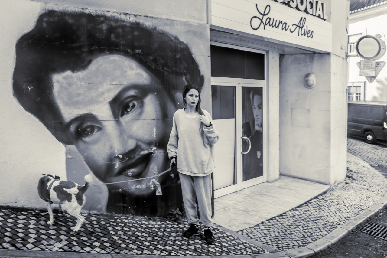 A woman and her dog on a lead on a street. The wall behind her has a mural with a woman's face, the actress Laura Alves. There's a doorway next to it and above the door is her name and just out of frame is the word "social".
