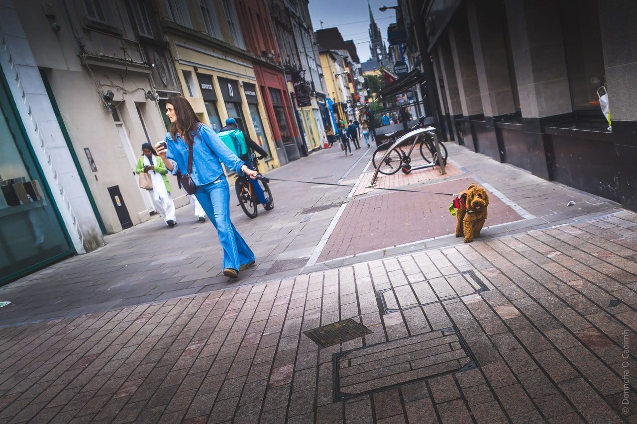 A woman with her walking along a street. She's carrying a coffee and her dog is on a lead, leaning away from her. The whole photo is tilted to the left, the opposite of the dog's tilt.