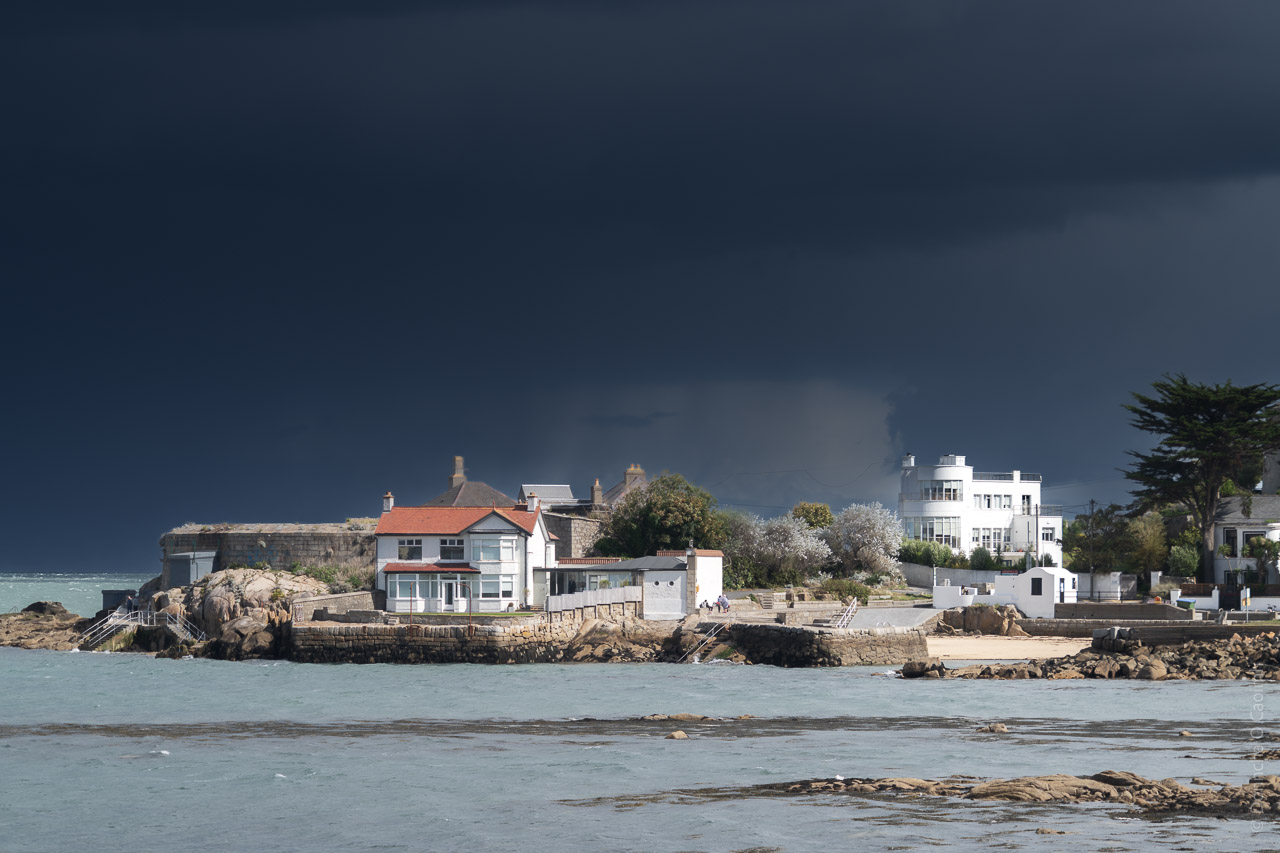 Dark clouds out at sea, beyond the pictured houses and coastline that are lit by the sun coming from behind me.