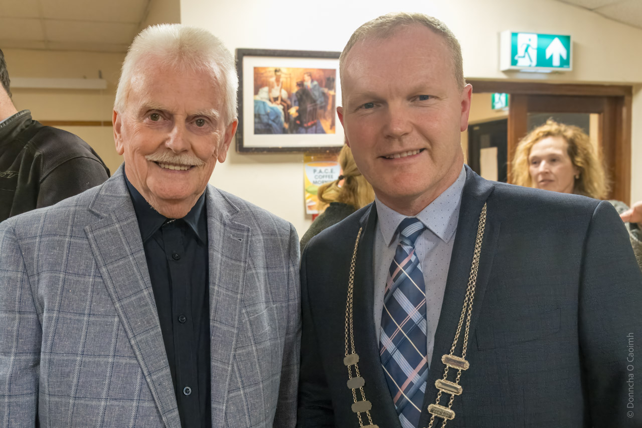 Joe Healy and Cllr. Seamus McGrath pose for a photo. Other people are visible in the background, as well as the door to the room, emergency exit sign and a painting on the wall.