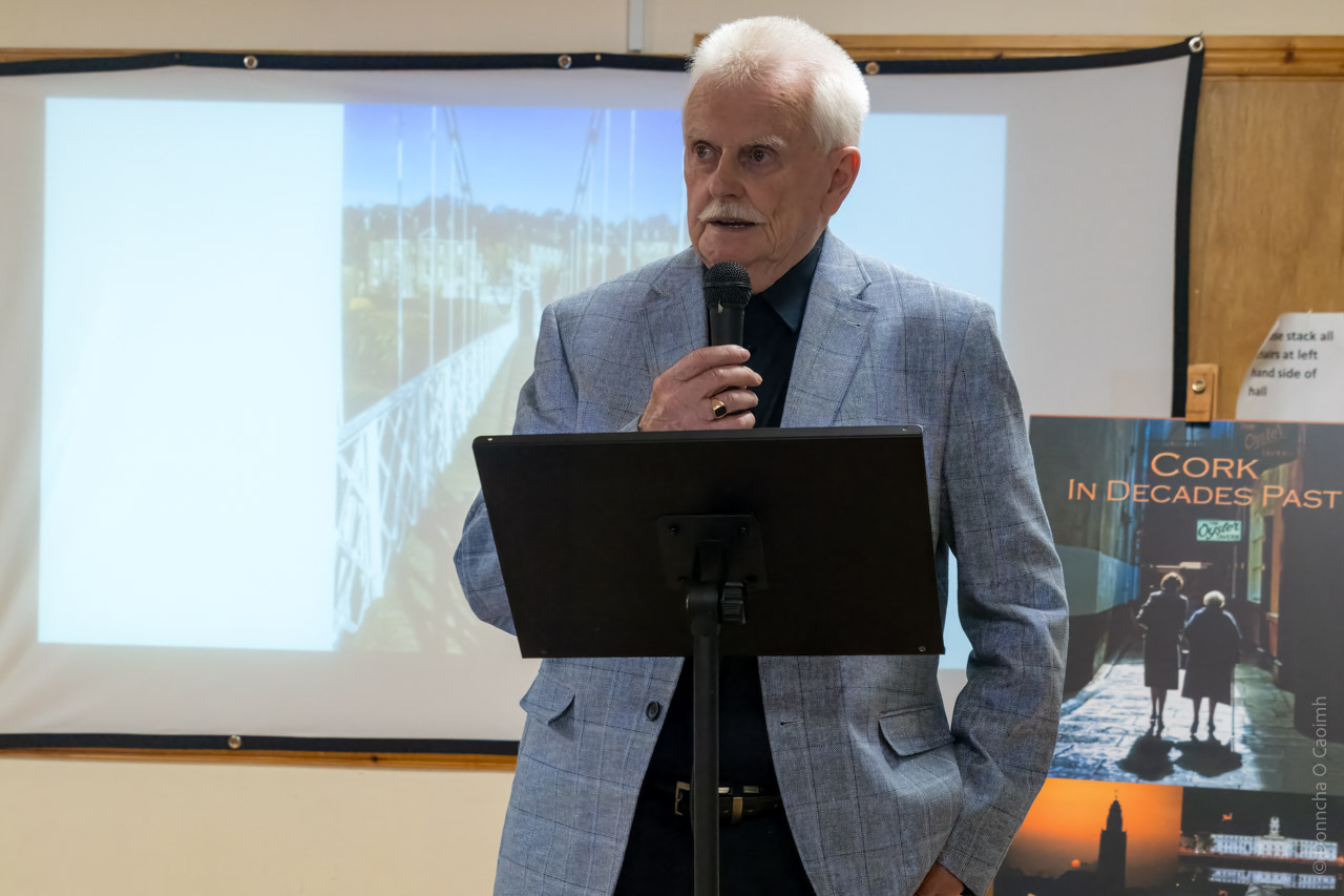 Joe Healy, speaking at a lectern into a microphone. A poster featuring his book cover is visible in the background, as is a projected image from the book.