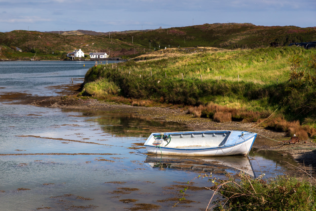 A blue and white boat is tied up on a gravel beach, with fields bordering the water area. Hills in the background and two houses as well.
