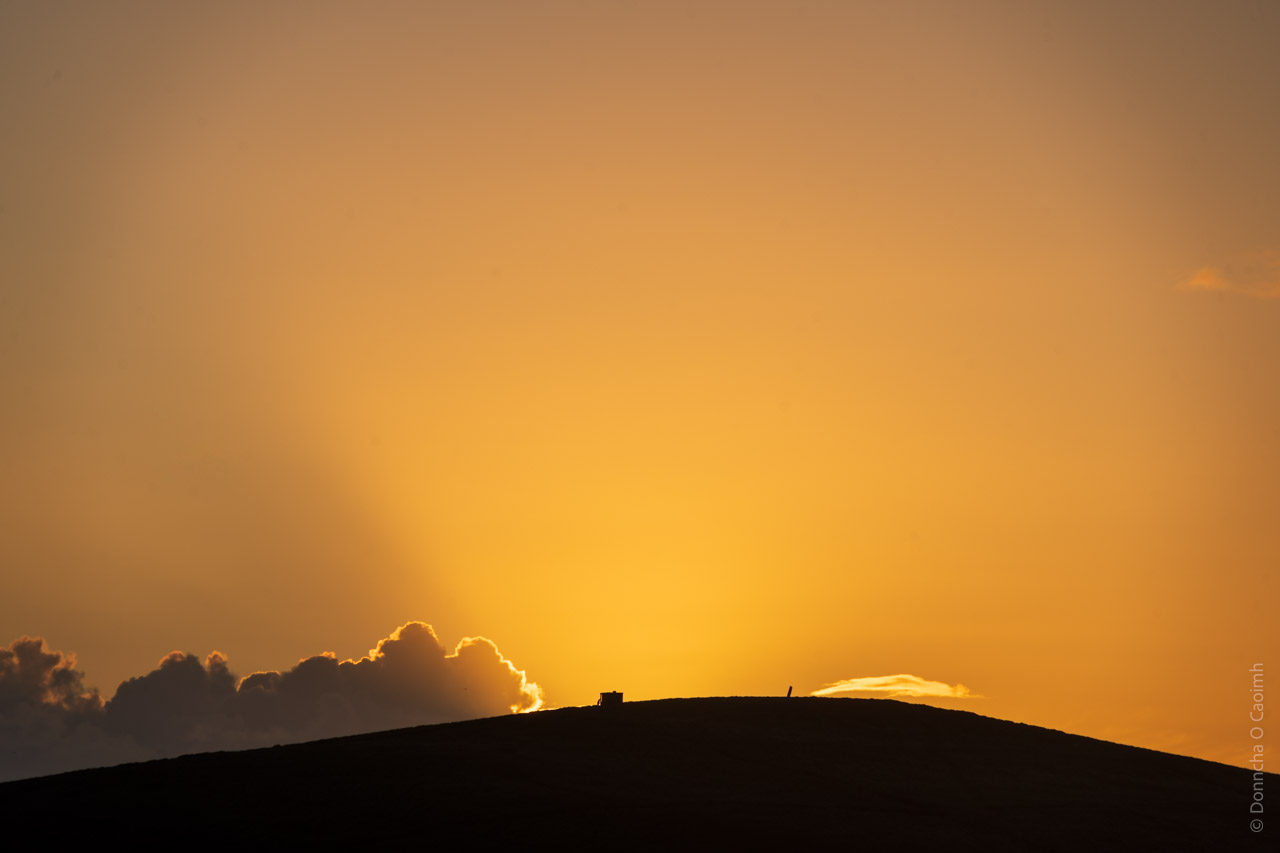 This image depicts a silhouette of a hill against a vibrant orange sky at sunset. Clouds are visible along the horizon, with sunlight creating a glowing outline around them. On the hilltop, there is a small structure and a vertical object, adding to the scene's serene atmosphere.