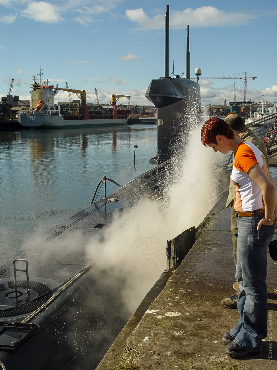The image shows a submarine docked at a port with steam or smoke being released from its side. In the foreground, a person with short red hair and wearing a white and orange shirt is standing on the dock, looking at the submarine. Another person is partially visible beside them. The background features a body of water with ships and cranes, indicating an industrial or harbor setting. The sky is clear with some clouds.