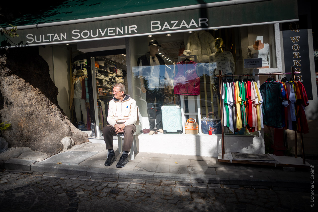 This image shows a shopfront of a "Sultan Souvenir Bazaar" with a person sitting outside on what appears to be a window ledge. The shop displays colorful clothing on racks, including t-shirts in various bright colors like green, yellow, and red. The storefront has a clean, modern appearance with large display windows showing mannequins and other merchandise. There's an interesting contrast between the modern shop front and what appears to be an old stone or rock wall formation to the left side of the store. The sidewalk is paved with cobblestones, giving it a traditional or historic feel. The lighting in the image creates an interesting effect, with natural sunlight casting shadows while illuminating the person sitting outside the shop. The overall composition suggests this might be located in a tourist area or historic district of a city.