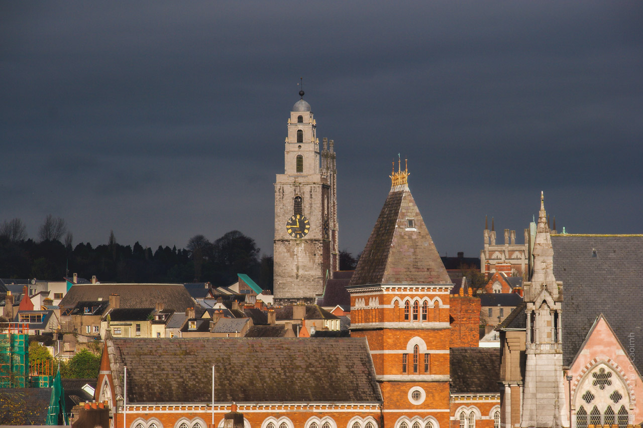 A dramatic cityscape of Cork, Ireland showing historic architecture against a dark, moody sky. The prominent Shandon Bells tower (St. Anne's Church) with its distinctive white facade and clock face dominates the skyline. The image features a mix of Victorian Gothic buildings with red brick facades, pointed spires, and traditional slate roofs. In the foreground are residential buildings and churches with characteristic architectural elements including arched windows, decorative stonework, and steep pitched roofs. The composition captures the layered nature of the city's historic center with buildings of varying heights and styles clustered together.