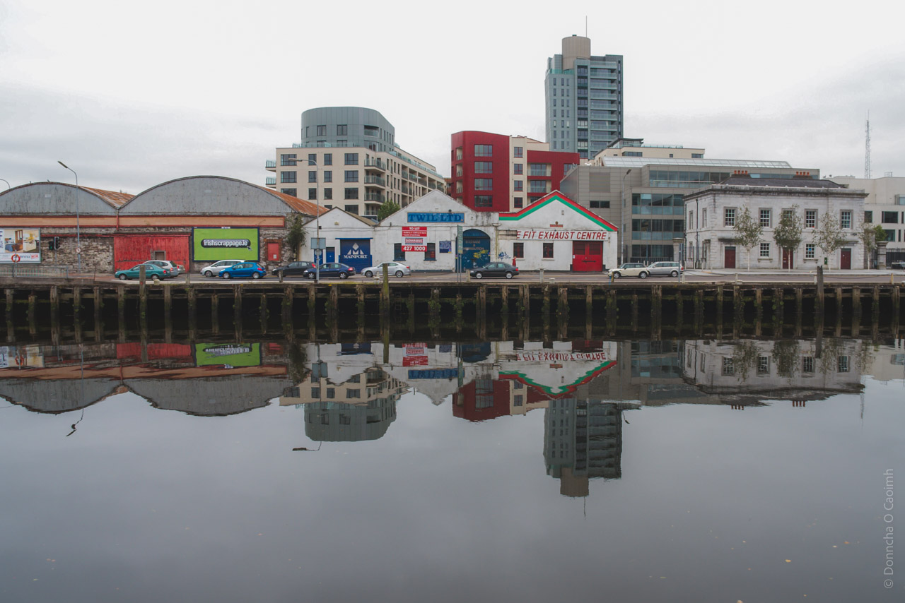 A cityscape reflection in Cork City, Ireland, showing a row of commercial buildings along the River Lee quayside. The scene features a mix of old warehouses with curved roofs, small businesses including a car exhaust center, and modern apartment buildings in the background. The still water creates a perfect mirror image of the buildings, with cars visible along the waterfront. The architecture contrasts between historic industrial structures and contemporary high-rise developments, captured under an overcast sky.