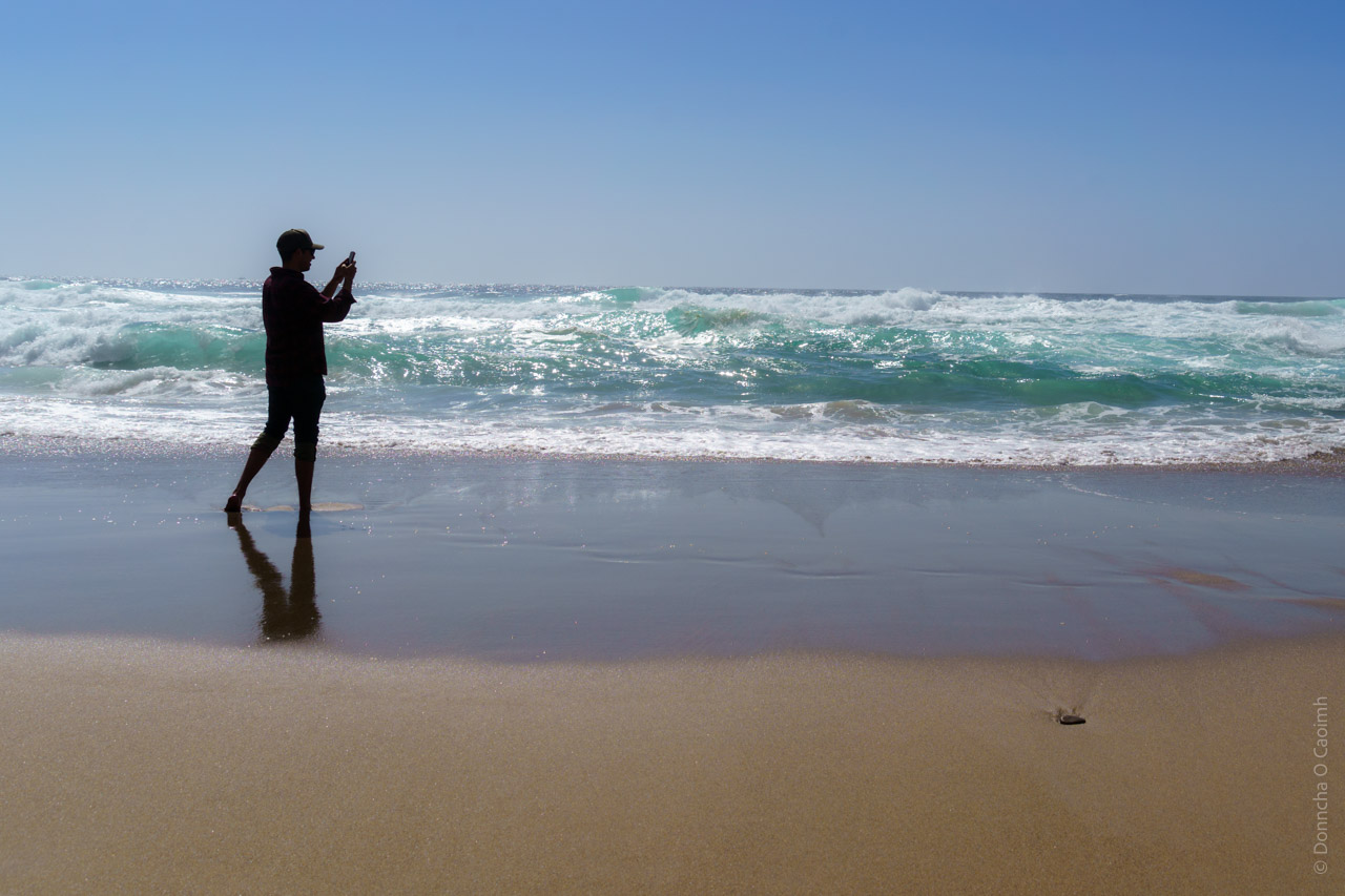 A silhouetted figure stands at the water's edge on a sandy beach, taking a photo with their phone. The person is wearing dark clothing and a cap. Turquoise waves crash in the background under a clear blue sky, creating a dynamic seascape. The wet sand reflects the figure's shadow, and the beach appears pristine with golden sand stretching into the frame.