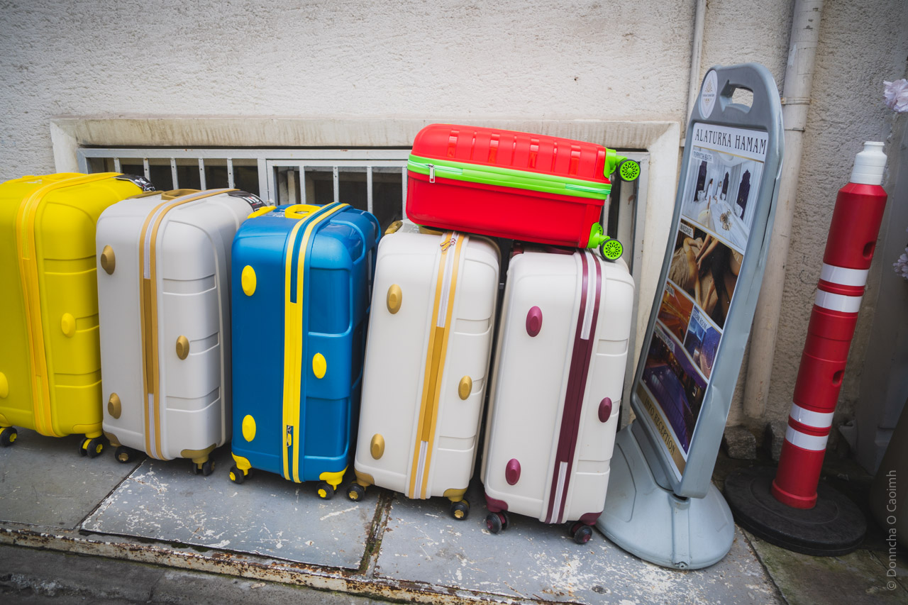A row of colorful hard-shell suitcases lined up against a white wall. From left to right: a yellow suitcase, two white suitcases with gold accents, a bright blue suitcase with yellow details, two more white suitcases with burgundy accents, and a red and green suitcase stacked on top. To the right of the luggage are two standing display signs, one labeled "Alaturka Hamam" and a red and white striped pole.
