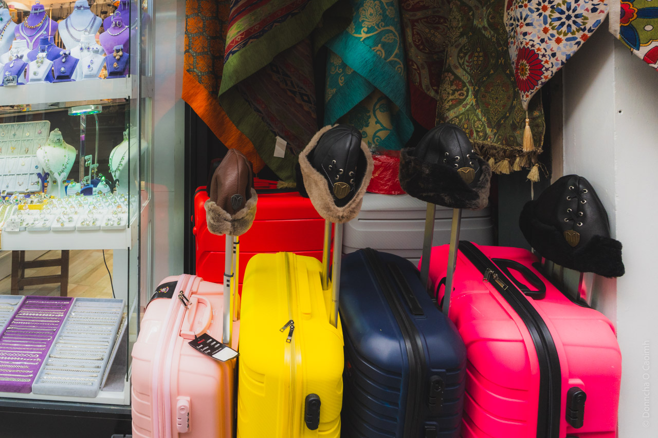 A retail display showing colorful hard-shell suitcases in pink, yellow, navy blue, and hot pink arranged in a row, with fur-trimmed winter hats displayed on stands above them. On the left side, there is a glass display case containing jewelry and necklaces, while decorative patterned fabrics or textiles in turquoise, orange, and green hang on the wall in the background.