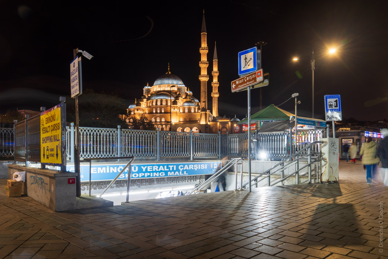 A nighttime view of the illuminated Yeni Camii (New Mosque) in Istanbul, with its distinctive Ottoman architecture featuring multiple domes and two tall minarets lit up in warm golden light. In the foreground, there's an entrance to an underground shopping mall called "Eminönü Yeralti Carsisi" with modern signage and handrails. The scene is framed by stone pavement and decorative metal railings, with street lights casting a glow on the surroundings. The contrast between the historic mosque and modern urban elements creates a striking composition typical of contemporary Istanbul's cityscape.