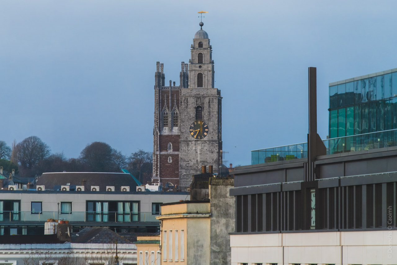 The iconic Shandon Bells tower of St. Anne's Church dominates the Cork city skyline, featuring its distinctive limestone construction with a golden clock face. The North Cathedral is visible behind it. Modern buildings, including glass and concrete structures, are visible in the foreground, creating a contrast between historic and contemporary architecture against a clear blue sky.