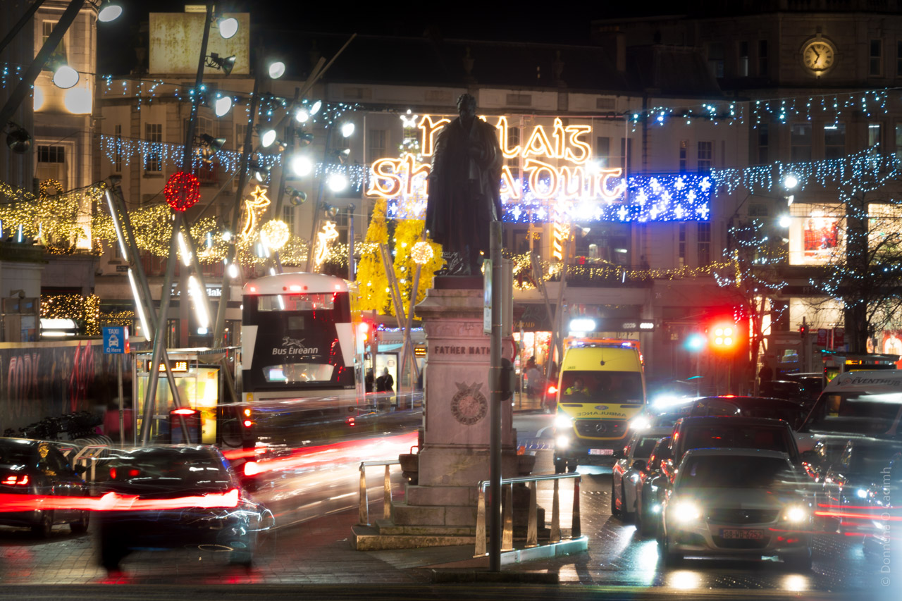 A nighttime street scene in Cork city centre during the Christmas season, showing the Father Mathew statue on Patrick Street illuminated by festive lights. The image features bright Christmas decorations including blue and gold string lights hanging overhead, with a prominent neon sign partially visible. Traffic is visible on the street, including a Bus Éireann bus and an ambulance with their lights on. The wet streets reflect the colourful holiday lights, creating a bustling, festive urban atmosphere.