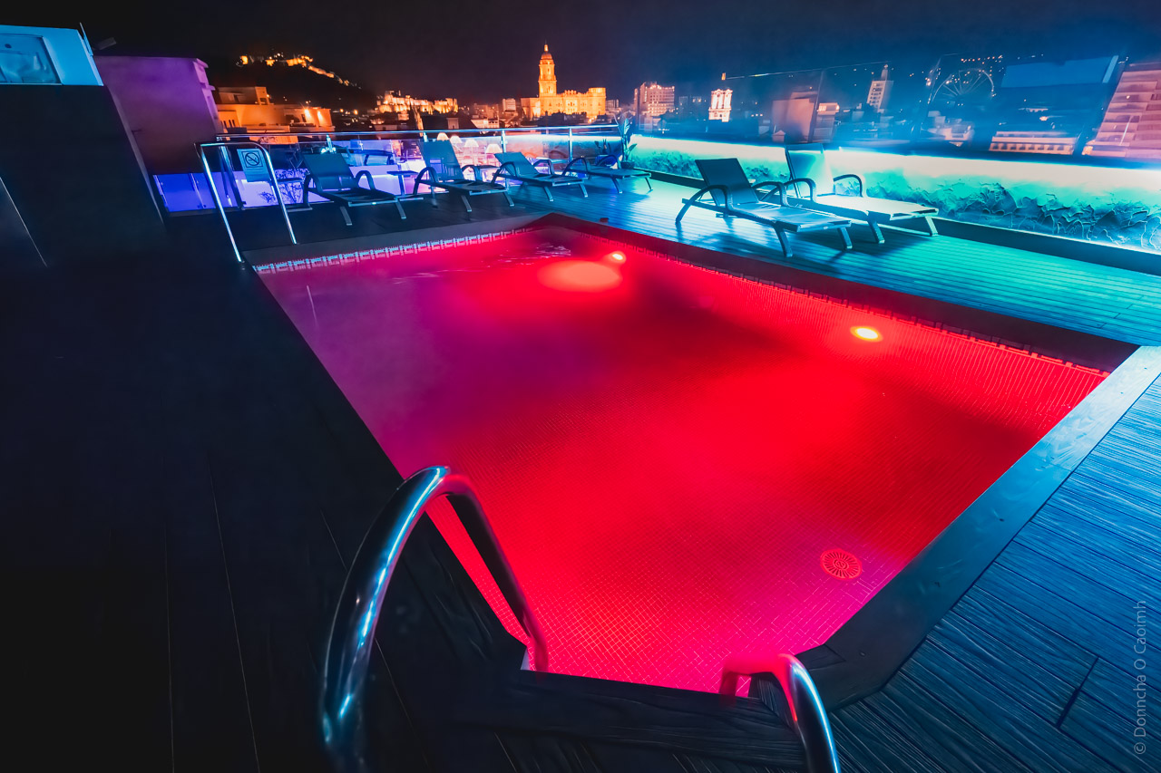 Rooftop pool at night in Malaga with red LED lighting illuminating the water, lounge chairs along the deck, and the illuminated cathedral visible in the city backdrop.