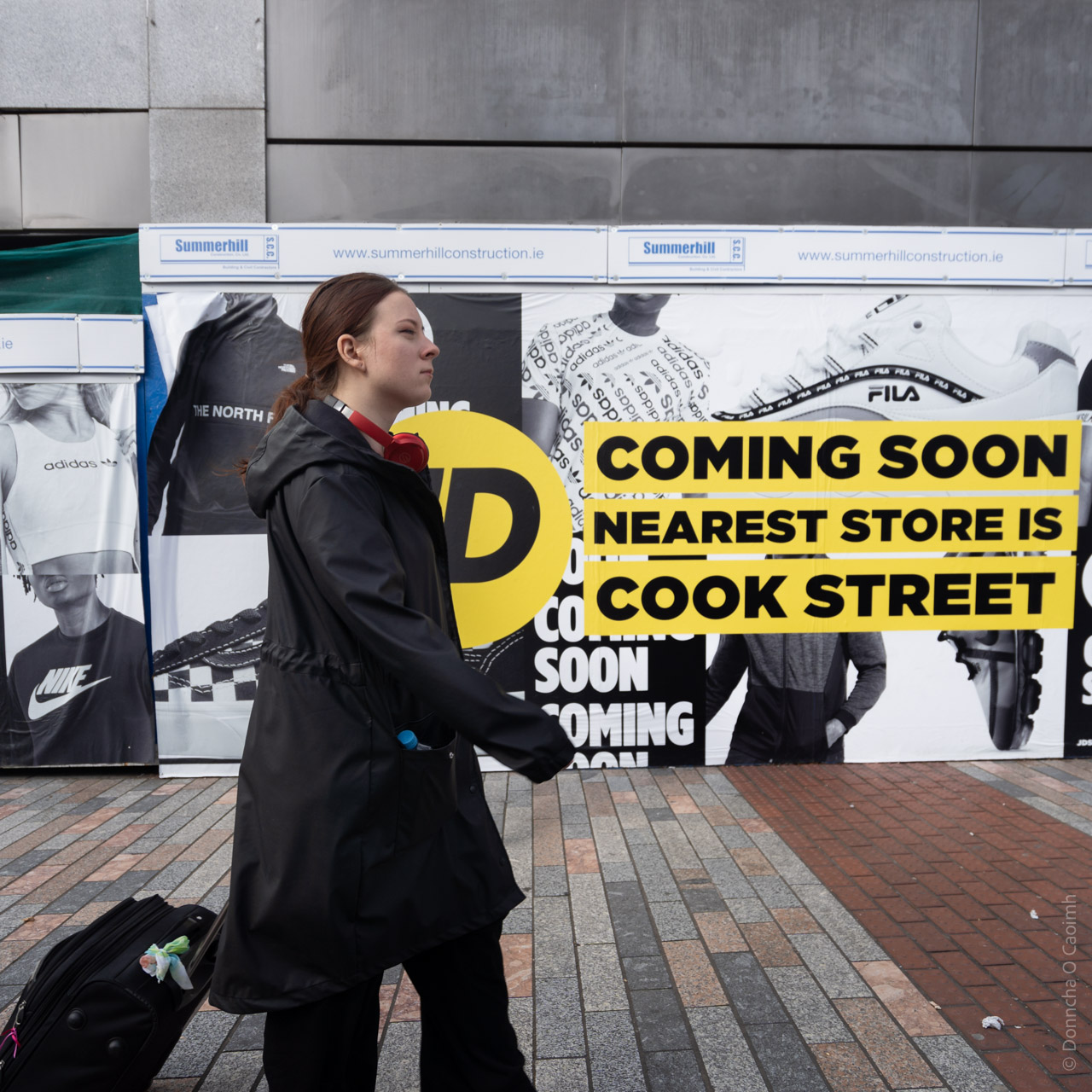 Person in a black coat walking past JD Sports construction hoarding with "COMING SOON" signage and Summerhill Construction branding, featuring sportswear advertising for Nike, Adidas, and Fila.