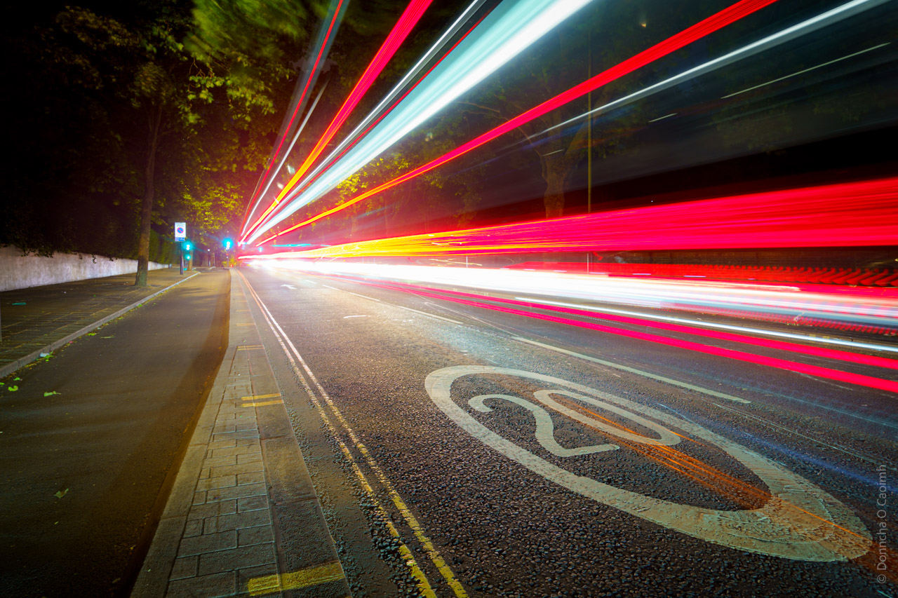 Long exposure night photograph of a city street showing streaking red and white light trails from passing traffic, with a "20" speed limit marking visible on the dark asphalt road surface, bordered by a pavement and trees in the background.