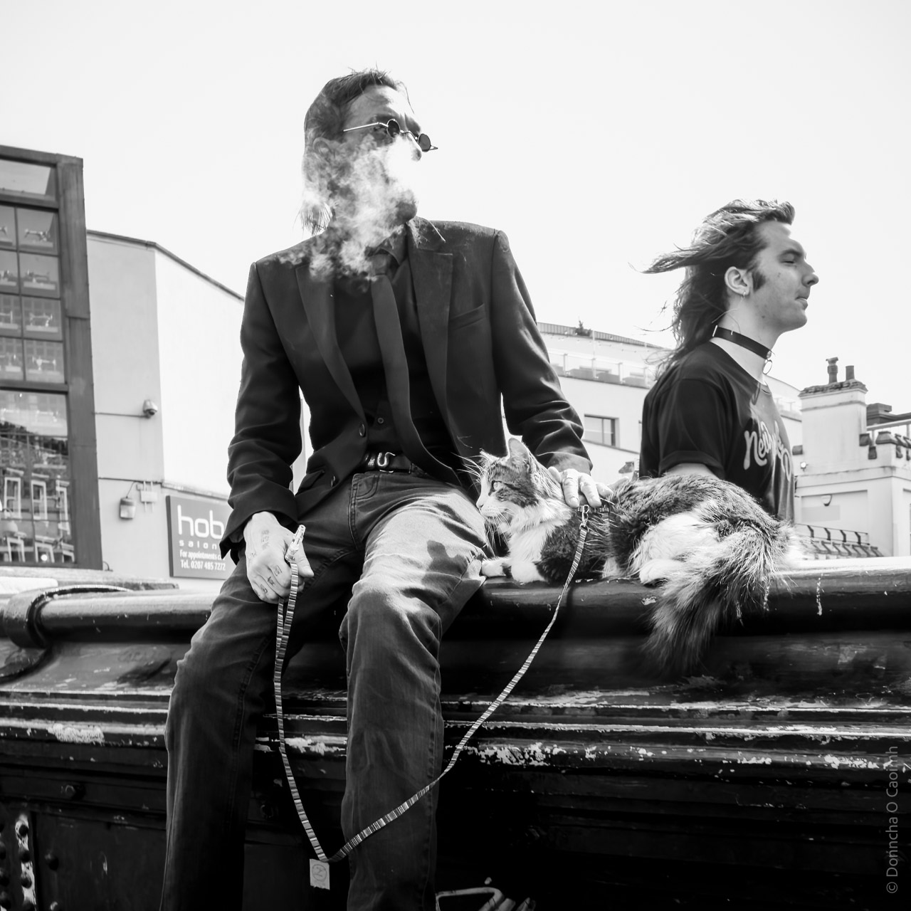A man in dark clothing sit on a ledge, smoking and wearing sunglasses while a leashed cat rests beside him. Another man walks past. An urban setting with "hob" signage visible in background.