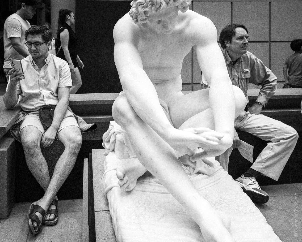 A classical white marble statue sits between two museum visitors on steps - one looking at a phone, another reading. The statue depicts a partially draped figure in a contemplative pose, while modern visitors appear absorbed in their own activities at the Musée d'Orsay.