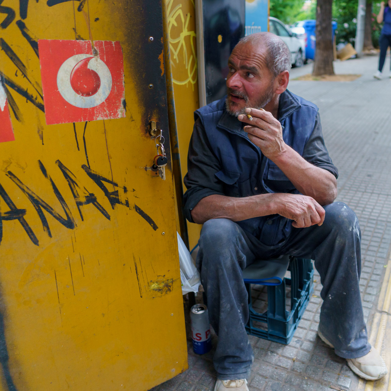 Street photograph: A middle-aged man with gray hair and beard sits on a blue plastic crate against a yellow wall with graffiti and a Vodafone logo, smoking a cigarette, wearing work clothes and appearing contemplative.
