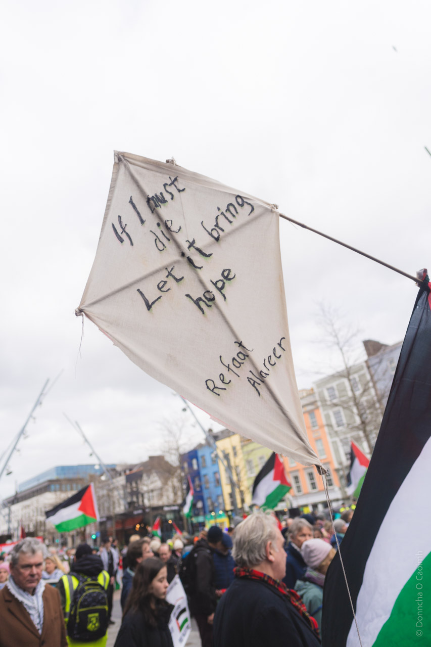 White kite with handwritten text "If I must die Let it bring hope" against urban backdrop with Palestinian flags visible.