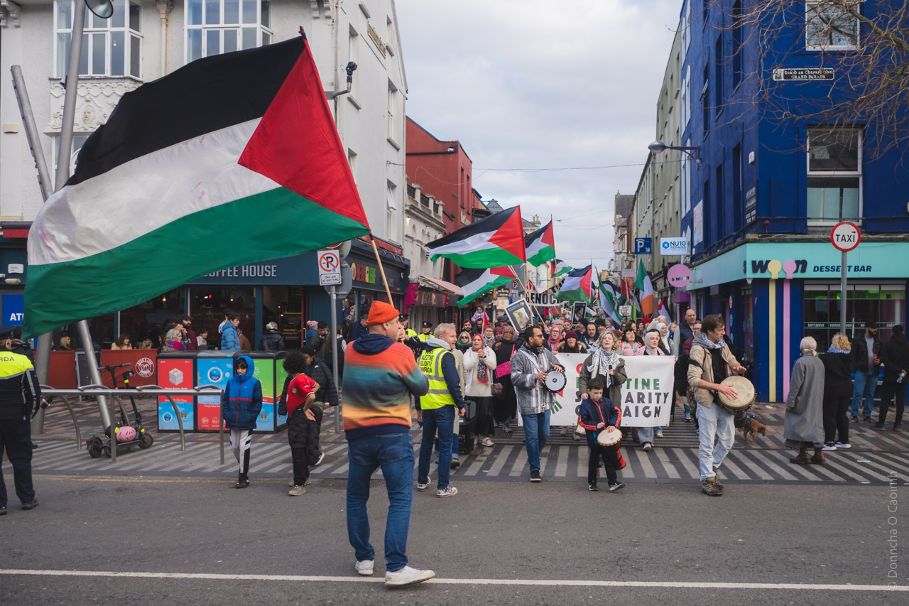 Pro-Palestinian march through Cork city streets with multiple Palestinian flags, protesters carrying drums and banners.