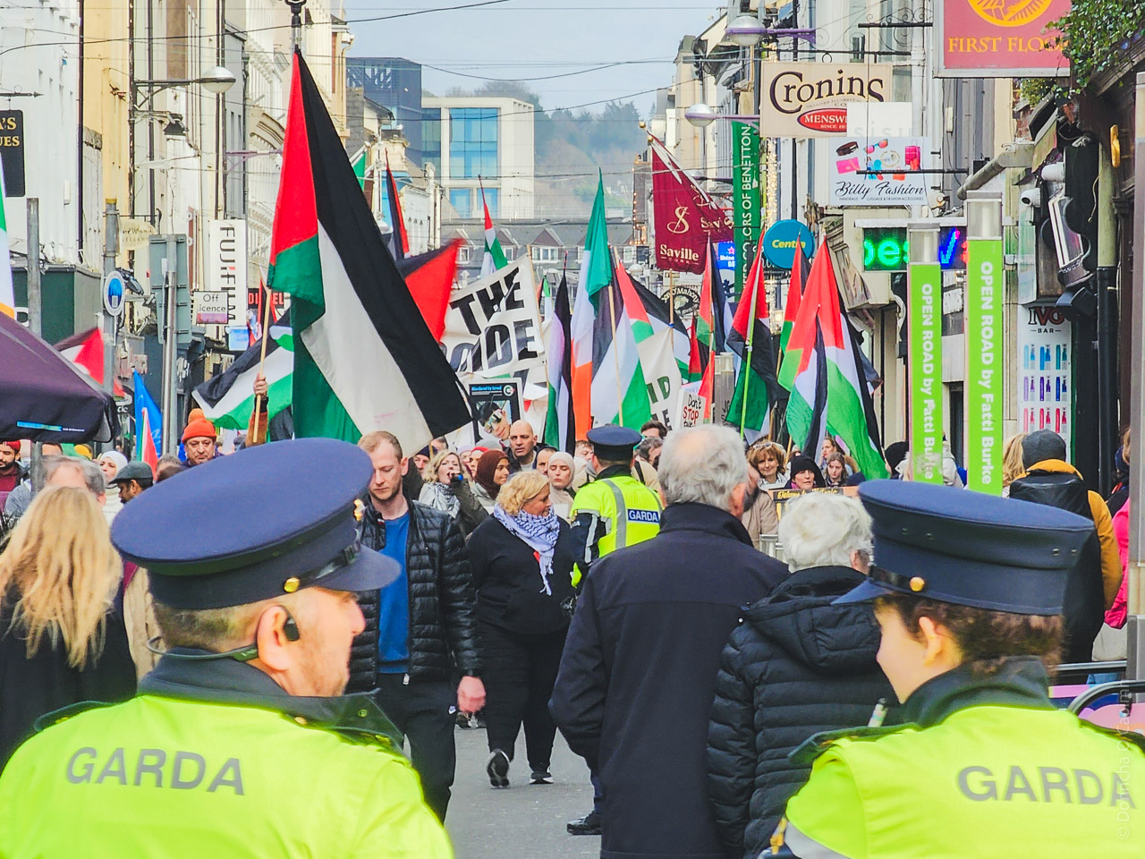 Pro-Palestinian protest in Cork city center showing large crowd with Palestinian flags and one Irish flag, with Garda officers monitoring the peaceful demonstration.