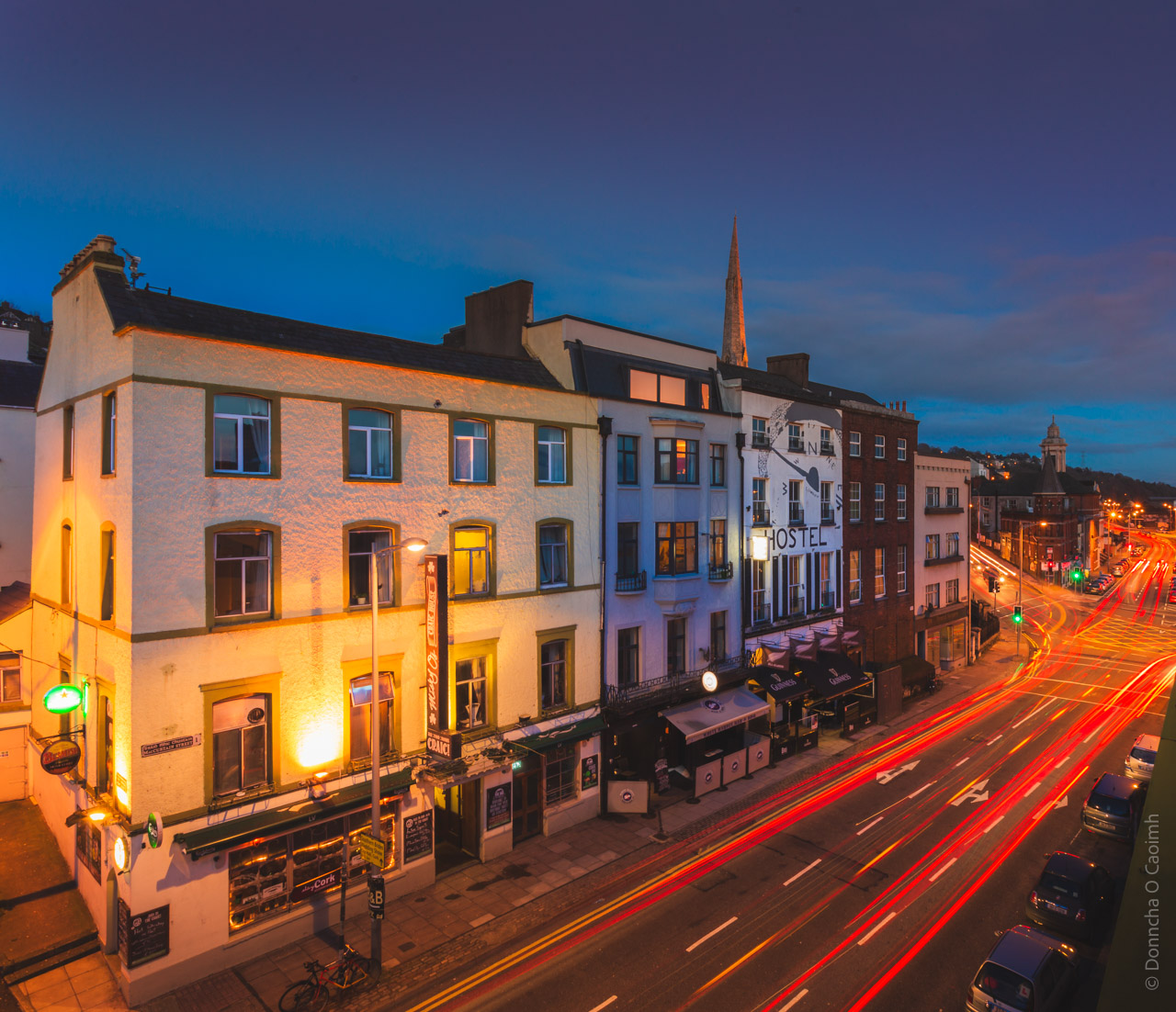 Night scene of McCurtain Street in Cork, Ireland showing illuminated Georgian buildings with a hostel, shops and pubs at twilight, with light trails from passing traffic creating red streaks along the road, and a church spire visible in the background.