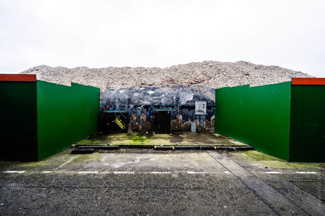 A symmetrical photo of a derelict building site entrance, flanked by green walls, with graffiti, moss and a pile of rubble behind, and the word "WESTCO" visible on a sign on the left.