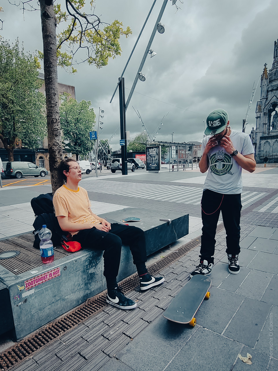 Two skateboarders in Cork City; one sitting on a concrete bench and the other standing, adjusting his headphones. The bench has a sticker that reads "ZOOTGHOST IS HOT."