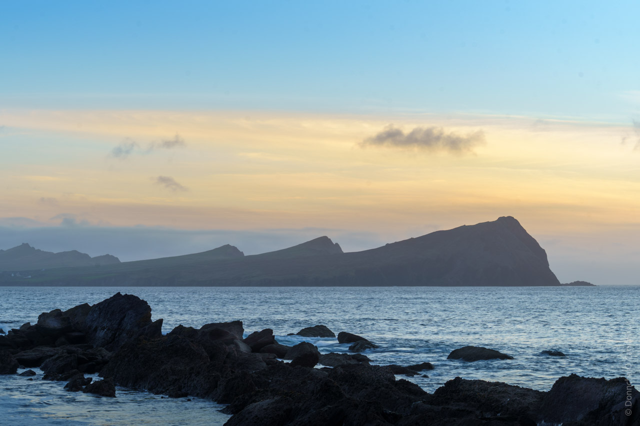 Sunset seascape showing the silhouetted peaks of Sybil Head and the Three Sisters on the Dingle Peninsula, with dark rocky foreground and golden sky reflected in the Atlantic waters.