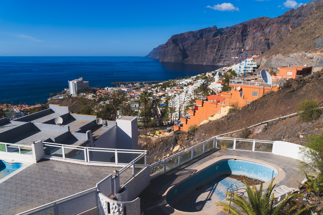 A view of Los Gigantes, Tenerife, showing dramatic cliffs, blue ocean, and residential buildings with swimming pools cascading down the hillside. Orange and white buildings dot the landscape against the backdrop of massive volcanic cliffs and deep blue Atlantic waters.