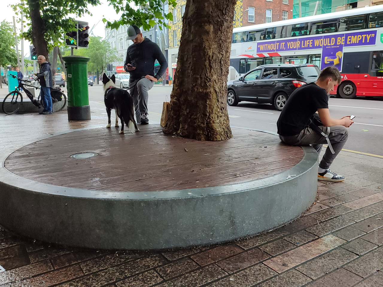 A street scene in Cork showing a round wooden bench around a tree with people using their mobile phones, a dog, a cyclist and a bus with the slogan "WITHOUT IT, YOUR LIFE WOULD BE EMPTY".