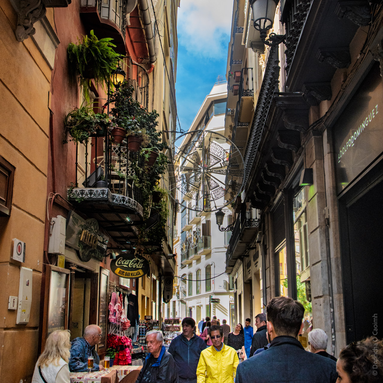 A vibrant street scene in Malaga, Spain, featuring a narrow alleyway with ornate balconies adorned with plants, a Coca-Cola sign for "Arte de Malaga," and outdoor seating with people enjoying drinks. A man in a bright yellow jacket stands out among the crowd, adding a pop of colour to the bustling atmosphere.