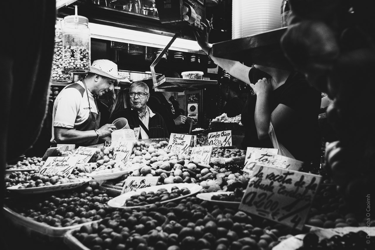 A black and white photo taken in Malaga, Spain, shows two men at a market stall piled high with olives and signs; one man in a hat serves a customer. The signs read "Gordal Piscante 0'250 /4", "Kimbo Chico 0'250 /4" and "la receta de la Abuela, corech 0'200/4".