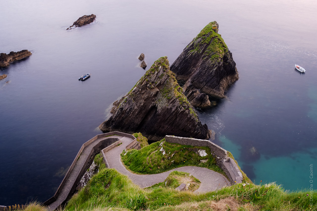 A scenic view of Dunquin Pier in County Kerry, Ireland, featuring winding stone paths leading down to the sea, triangular rocky outcrops covered with green grass, and small boats floating in calm waters.