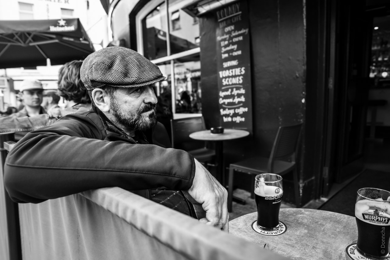 A black-and-white photograph of a man wearing a flat cap and jacket, sitting outdoors at a pub table in Cork, Ireland. He leans on a barrier, with two pints of Murphy's stout on the table in front of him. A chalkboard menu behind him lists items such as "Toasties" and "Scones" alongside coffee options. Other patrons are visible in the background, seated under umbrellas.