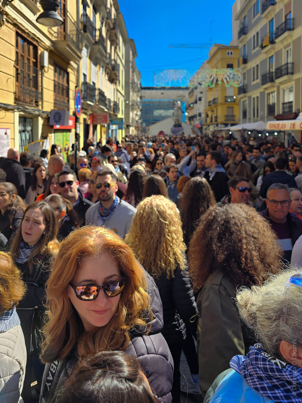 A vibrant street scene in Valencia, Spain, during the Fallas festival. The street is crowded with people enjoying the festivities, surrounded by historic buildings and decorations. A statue is visible in the background, and colourful banners hang across the street under a clear blue sky. 