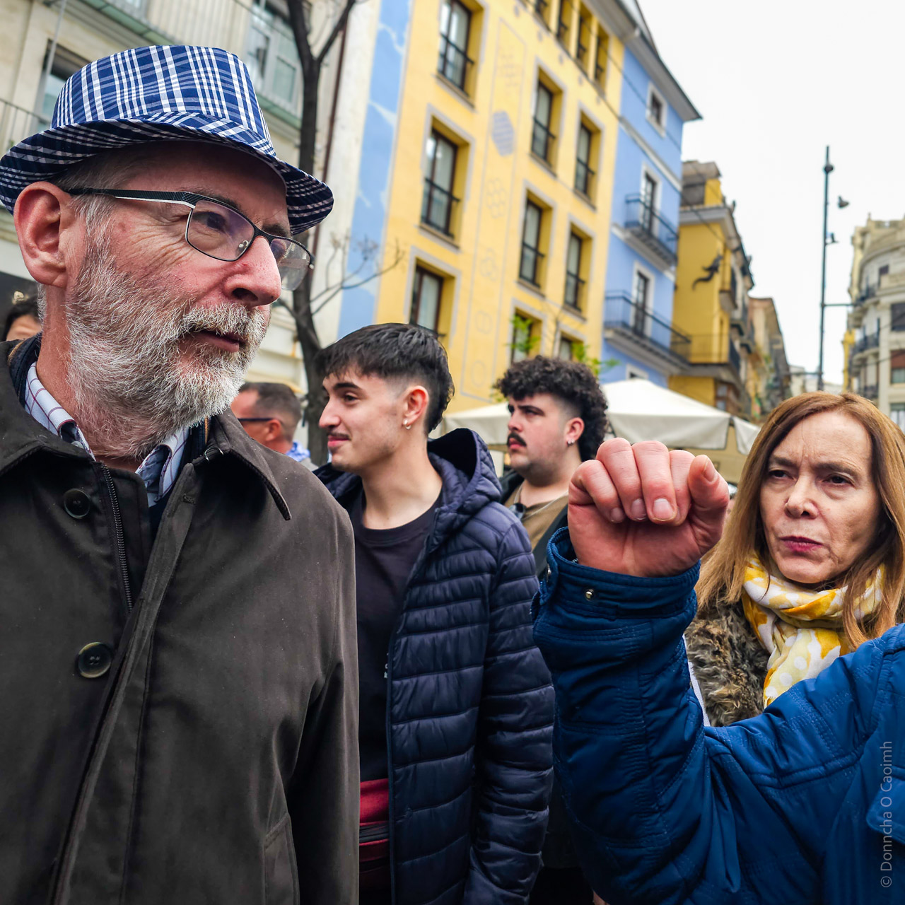 A bustling street scene in Valencia, Spain, featuring a group of people in casual clothing surrounded by colourful buildings with intricate facades. A man wearing a plaid hat and brown jacket is in the foreground, while another person raises their hand in a gesture. The background includes vibrant blue and yellow architecture and street activity.