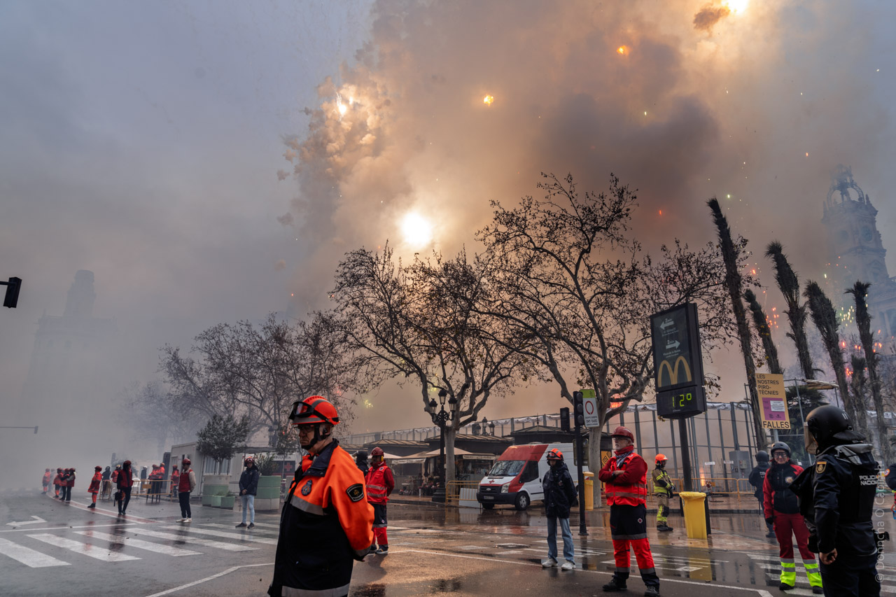 Smoke fills the sky during a pyrotechnic event in Valencia, Spain's Town Hall Square. Flashes of light from fireworks light up the smoke. Emergency workers in bright uniforms stand in the foreground as the spectacle unfolds against a backdrop of historic architecture, including the clock tower of the Town Hall. Signs for McDonald's and "FALLES" are visible on the street.
