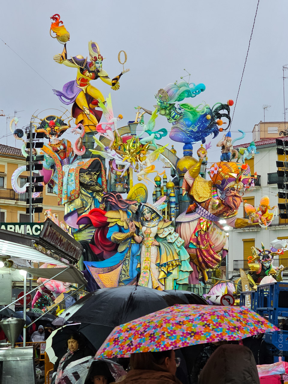 A vibrant and intricate Fallas sculpture in Valencia, Spain, depicting colourful figures in elaborate costumes, including a woman in a detailed dress, mythical characters, and fantastical elements. The scene is set on a rainy day with umbrellas in the foreground and buildings in the background.