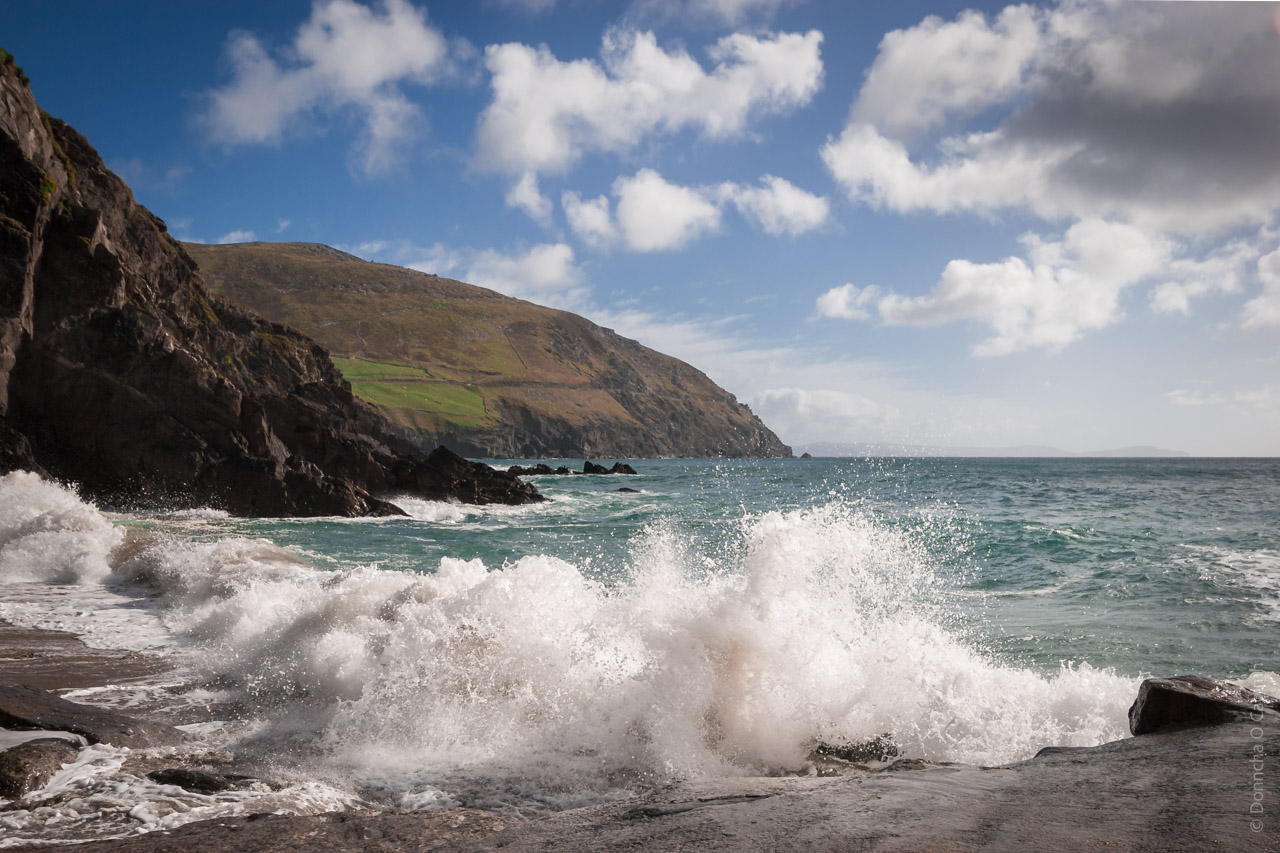 A dramatic coastal scene featuring waves crashing against rocky cliffs under a bright blue sky with scattered clouds, with lush green hills in the background.