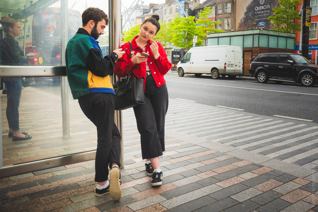 A candid street photo taken on Grand Parade, Cork, Ireland in 2018, showing a young man and woman at a bus stop, with their phones out, and a another woman in the background, city traffic and buildings in the background, and a billboard reading "QUERCUS Scholarship" visible.